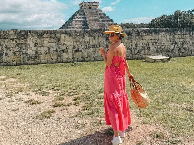 Eine Frau in einem rosa Kleid geht bei Chichén Itzá an Steinmauern entlang.